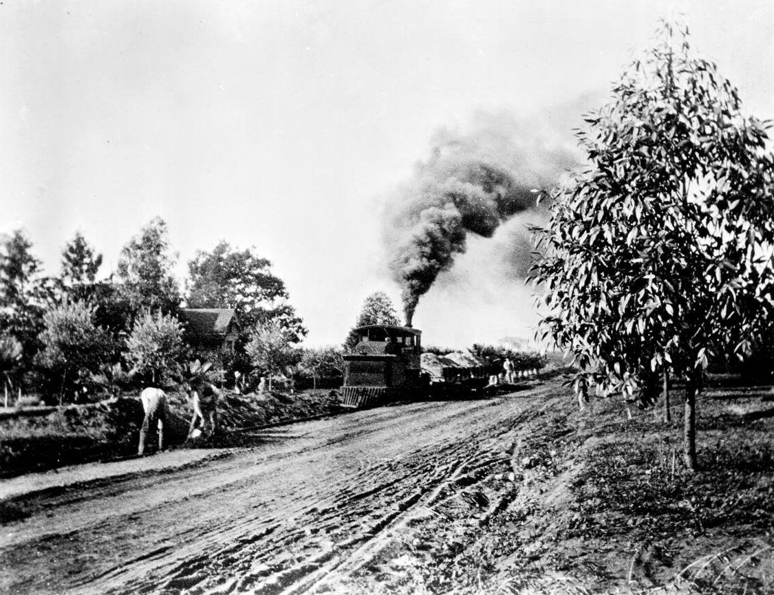 A steam train hauling dirt along an unpaved road while workers dig beside it